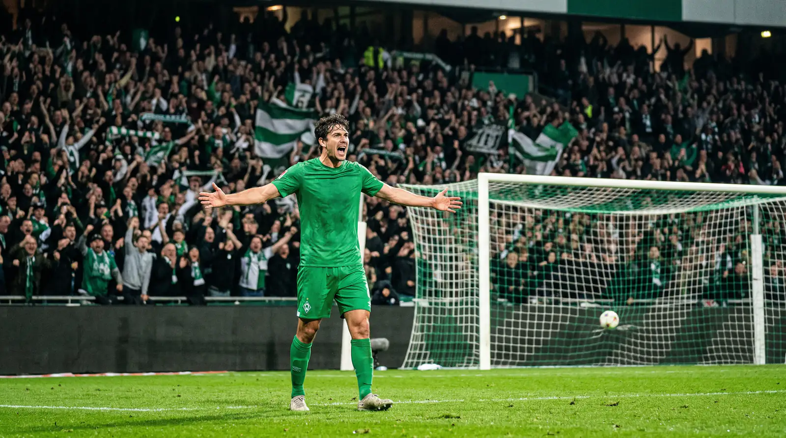 Fußballspieler feiert sein Tor mit ausgebreiteten Armen vor der Fankurve